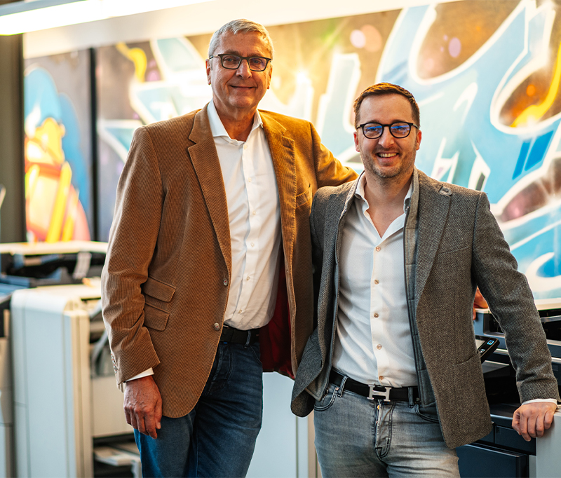 Deux managers souriants, debout côte à côte, représentant la direction de D&O Partners, forte de plus de vingt ans de confiance et de partenariat avec Xerox, devant un fond artistique du Showroom de D&O Partners.